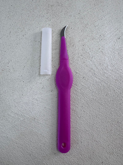 A pink seam ripper by Super Cheap Fabrics rests on a textured gray surface, featuring a precision curved blade and a long white cap.