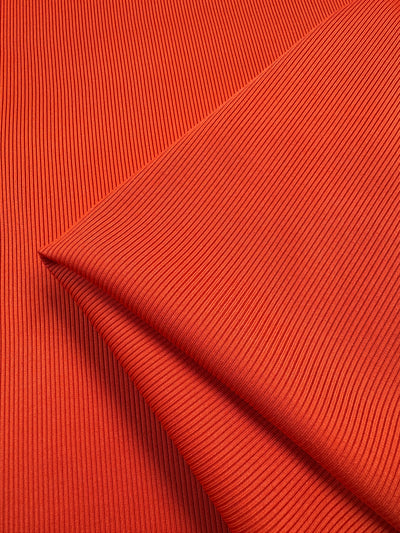 The close-up of Super Cheap Fabrics' Rib Knit 1x1 in Tango showcases vibrant orange pleats, creating a textured pattern with a medium weight feel. The fabric's reflective surface and stretch create dynamic movement and subtle hue variations. Width: 127cm.