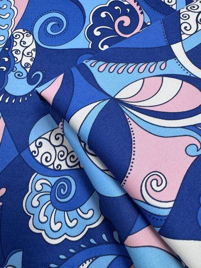 A close-up of Super Cheap Fabrics’ Cotton Sateen – Serenity Waves (148cm), a lightweight fabric with bold, abstract swirls, paisleys, and curved lines in blue, light blue, pink, and white. The sateen is shown slightly folded.