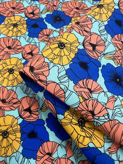 A close-up of vibrant fabric adorned with a floral pattern. The medium to heavy weight linen-cotton blend features yellow, blue, and pink flowers with black centers against a light teal background. The flowers overlap and are densely arranged, creating a lively, colorful design perfect for home décor.
Product Name: Printed Canvas - Wonderland - 145cm  
Brand Name: Super Cheap Fabrics