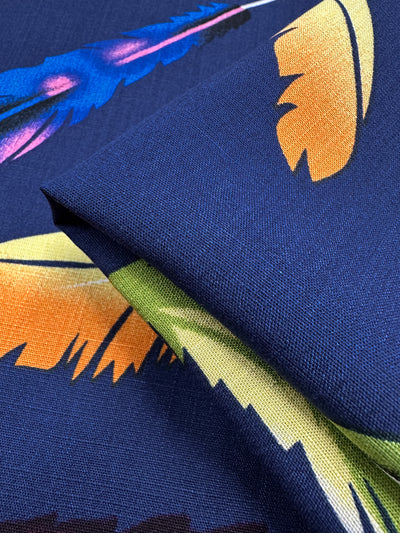 Close-up of Super Cheap Fabrics’ Bamboo Rayon - Feather Blue (150cm): lightweight, dark blue fabric with vibrant yellow, orange, green, and blue feather prints. Neatly folded to display texture—ideal for beginner sewing projects.