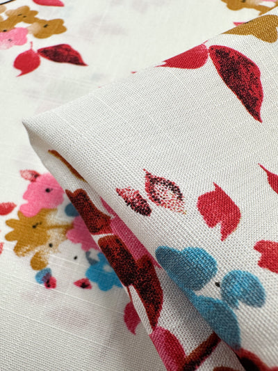 A close-up of Super Cheap Fabrics’ Bamboo Rayon - Lil Flower (150cm) shows its lightweight white fabric with a vibrant floral print in red, gold, pink, and blue. The partially folded material highlights its soft texture and colorful design.