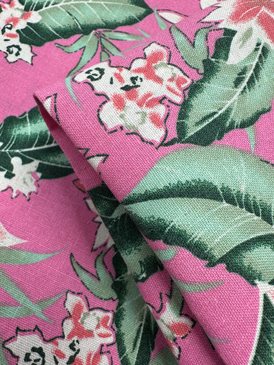 Close-up of Super Cheap Fabrics' Printed Linen Blend - Jungle Bloom (140cm), showing pink fabric with a tropical floral pattern—large green leaves and white-pink flowers. The medium weight linen cotton blend is partially folded for added texture.