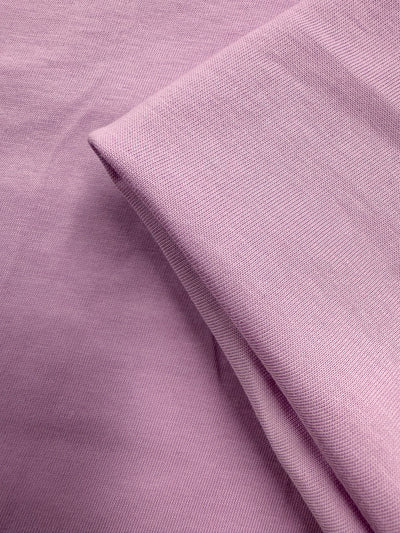 A close-up of Super Cheap Fabrics' Cotton Jersey - Orchid Bouquet (165cm) shows one section folded over another, highlighting the soft texture, smooth weave, and light to medium weight of this purple fabric.