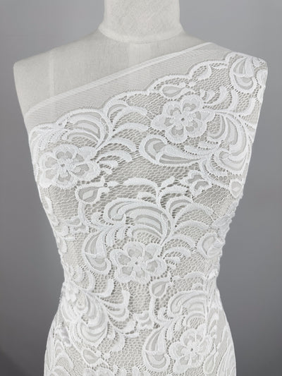 A close-up of Designer Lace - White - 145cm by Super Cheap Fabrics on a one-shoulder dress form showcases its floral and swirl patterns, ideal for elegant evening dresses. The neutral gray background accentuates the intricate texture and detail of the fabric.