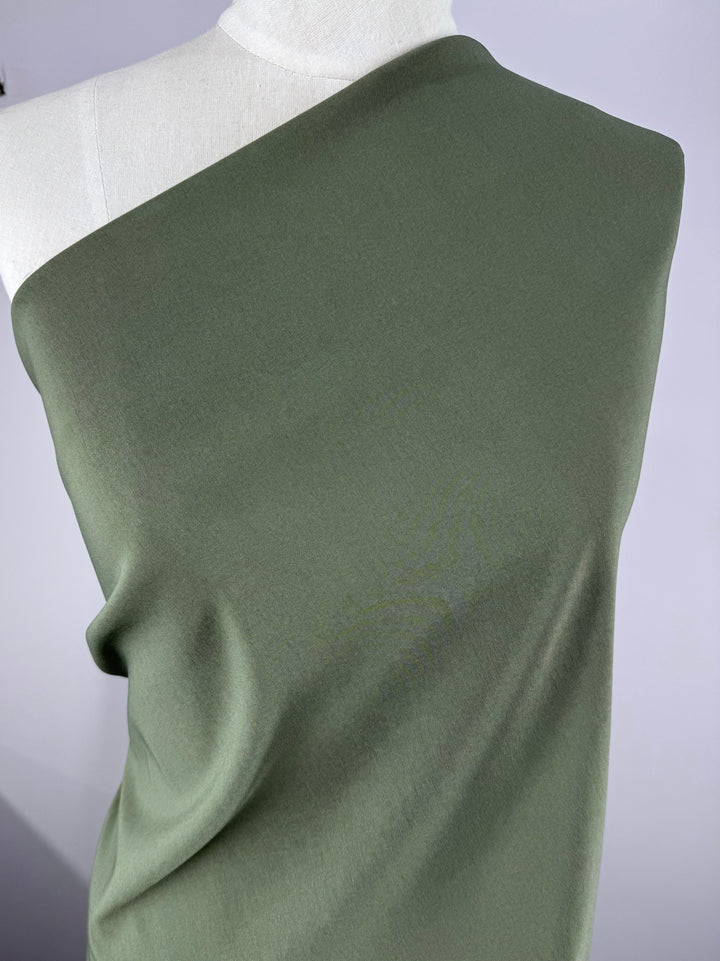 A mannequin gracefully displays the Stretch Bengaline - Beetle (150cm) by Super Cheap Fabrics, styled in an off-shoulder olive-green design. The plain white background enhances the elegance and flow of this spandex blend.