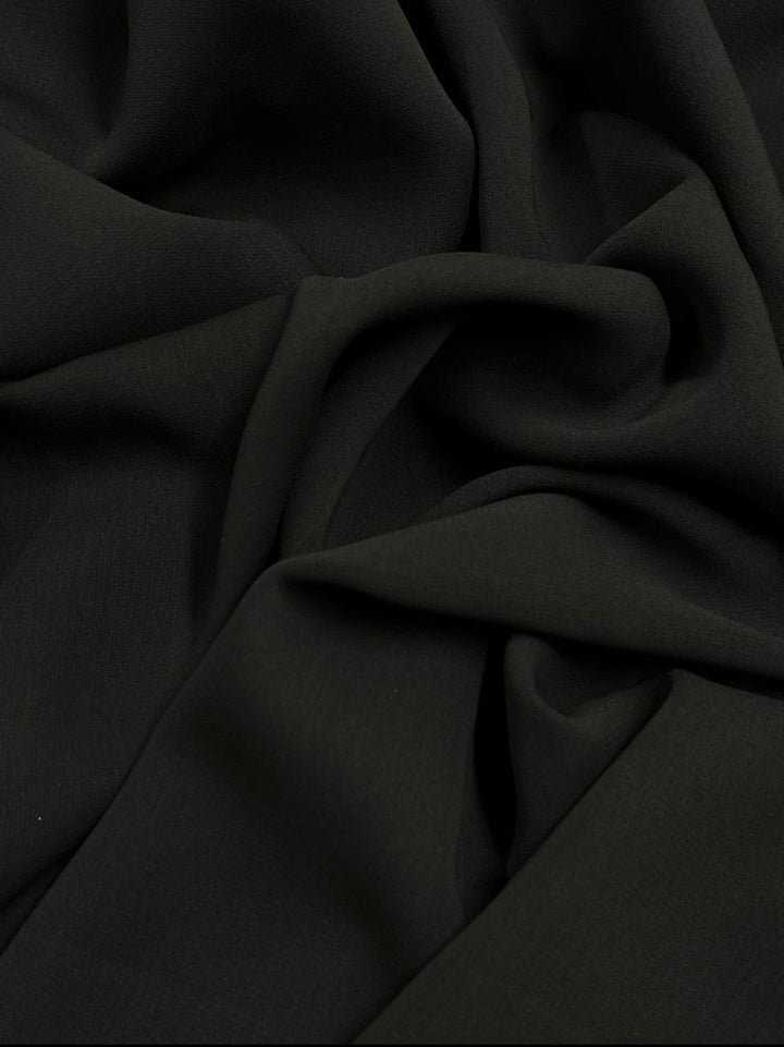 The Stretch Crepe - Raven 130cm by Super Cheap Fabrics features a dark green crepe fabric, showcasing smooth, flowing texture with gentle folds and creases, enhanced by a spandex blend for added stretch.