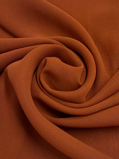 Close-up of Hi-Multi Chiffon in Rust by Super Cheap Fabrics (148cm), lightweight fabric neatly folded in a spiral, highlighting its smooth texture and subtle ribbed detail.