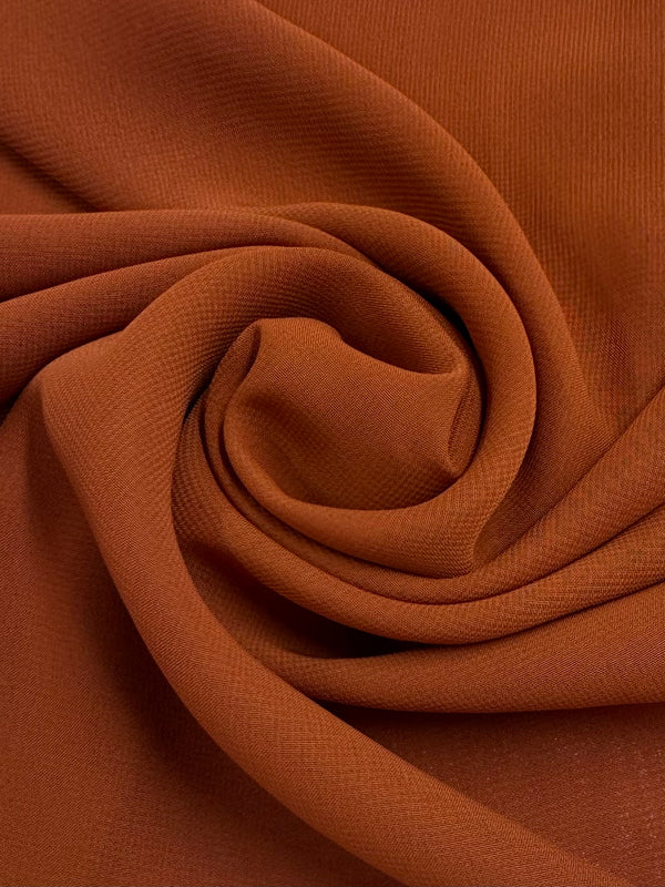 Close-up of Hi-Multi Chiffon in Rust by Super Cheap Fabrics (148cm), lightweight fabric neatly folded in a spiral, highlighting its smooth texture and subtle ribbed detail.