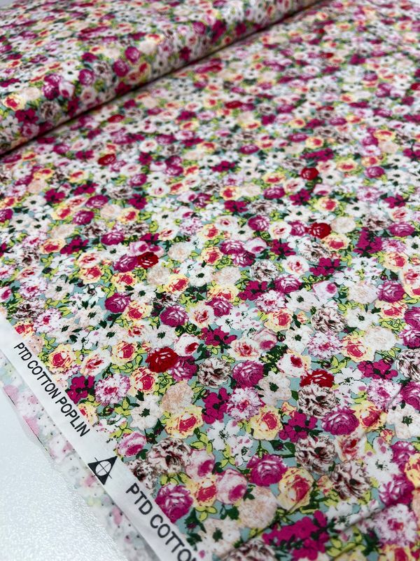 Close-up of the Printed Cotton - Roseness - 120cm by Super Cheap Fabrics, showcasing natural cotton fibers. This fabric displays a vibrant blend of red, pink, yellow, and white flowers against a white backdrop, making it versatile for multiple projects. The edge is marked with "PTD COTTON POPLIN.