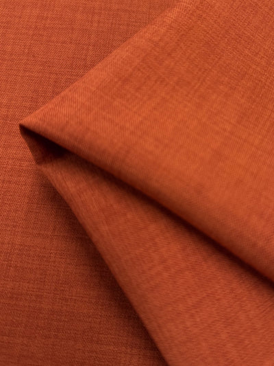 A close-up of Twill Suiting - Rust - 150cm by Super Cheap Fabrics shows folded burnt orange polyester with a subtle, fine texture and intersecting diagonal lines, highlighting its soft twill weave.