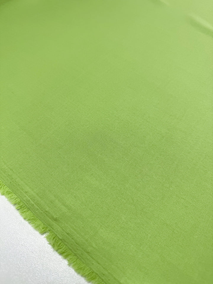 A close-up of Super Cheap Fabrics' Twill Suiting - Green Glow (150cm), showing smooth, light green polyester fabric with a diagonal twill weave and a slightly frayed edge, laid flat on a white surface.