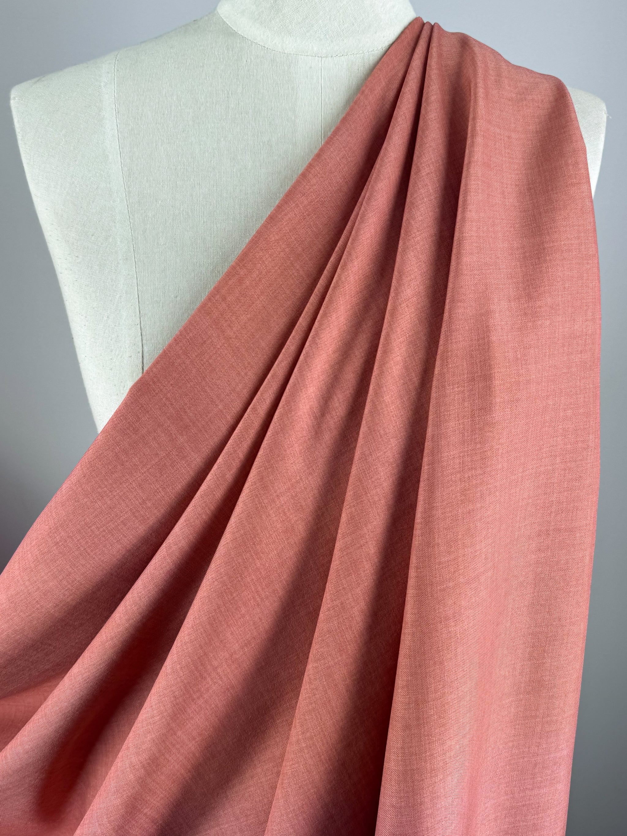 Super Cheap Fabrics’ Twill Suiting - Terra Cotta (150cm), a 100% polyester fabric with a pinkish-coral hue and diagonal weave, is draped over a cream dress form, showcasing its soft, elegant folds against a neutral background.