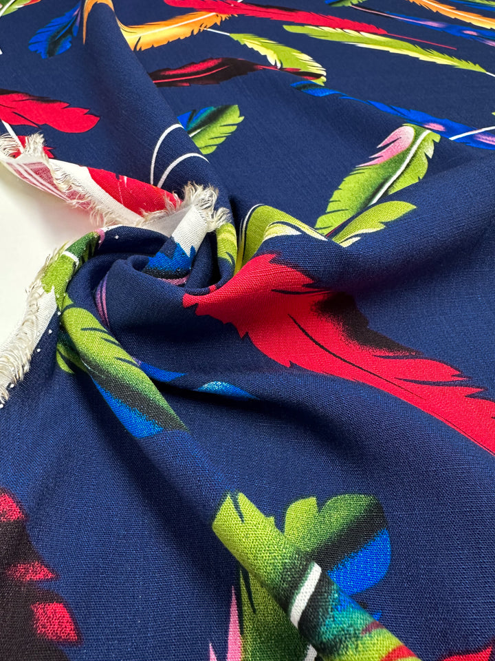 A close-up of Super Cheap Fabrics’ Bamboo Rayon - Feather Blue - 150cm shows lightweight blue fabric with red, green, yellow, and pink feather prints. It’s twisted at the center, with a frayed edge on the left.