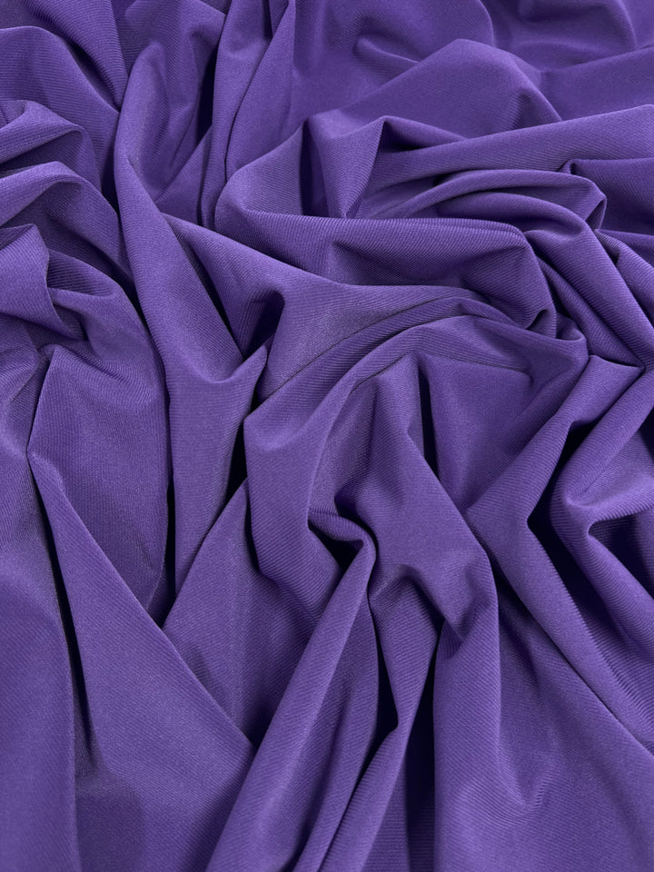 Crinkled, deep purple fabric with soft folds creating an intricate pattern. The texture appears smooth and slightly shiny, reflecting light gently. This medium weight fabric is ITY Knit - Pop Purple - 150cm by Super Cheap Fabrics, offering a pop of purple that's both vibrant and durable.