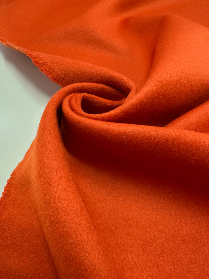 A close-up of Super Cheap Fabrics’ Wool Cashmere - Koi - 150cm, showing its vivid orange hue, luxurious softness, smooth texture, and a softly swirled fold with an unfinished edge.
