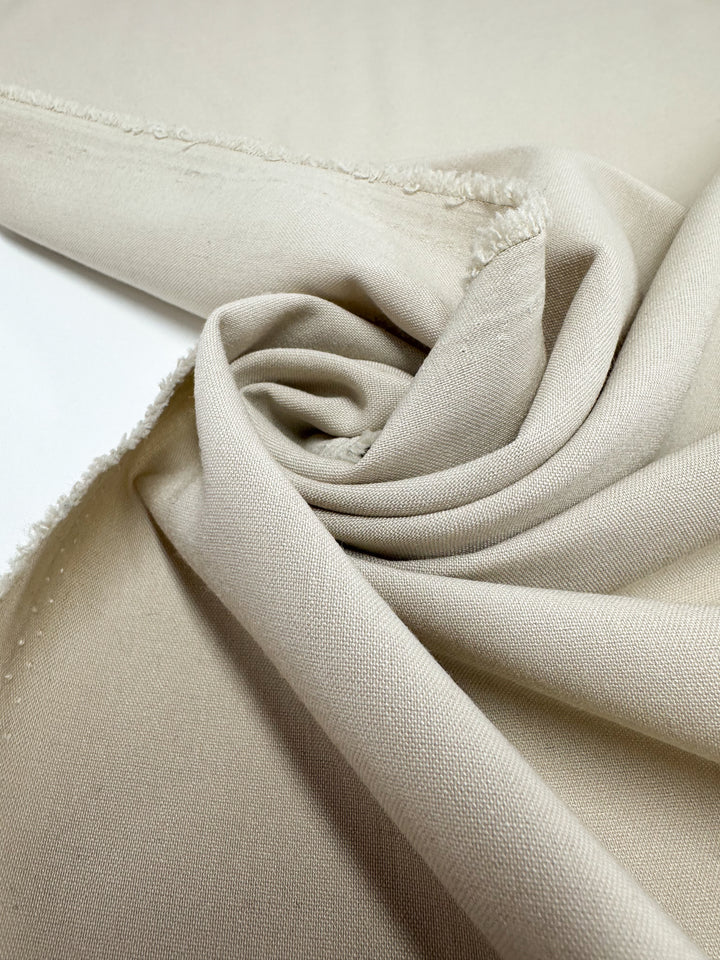 A close-up of Suiting - Oatmeal - 150cm by Super Cheap Fabrics, showcasing its polyester viscose blend, slightly rough texture, weave details, and unfinished edges as it is partially rolled and loosely folded.