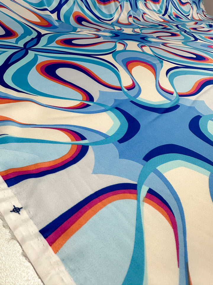 A close-up of Silky Satin - Burst - 150cm by Super Cheap Fabrics showcases vibrant blue, red, and orange abstract swirls on white satin—creating a dynamic, retro-inspired look ideal for elegant garments or printed satin projects.