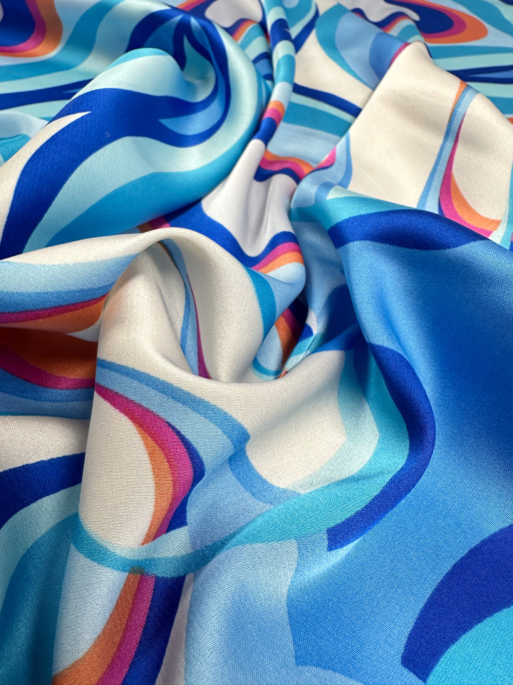 Close-up of Silky Satin - Burst, a Super Cheap Fabrics product, showcasing a vibrant abstract pattern with swirls in blue, teal, pink, and orange. The lightweight 150cm satin has a smooth texture that captures light for gentle highlights and shadows.