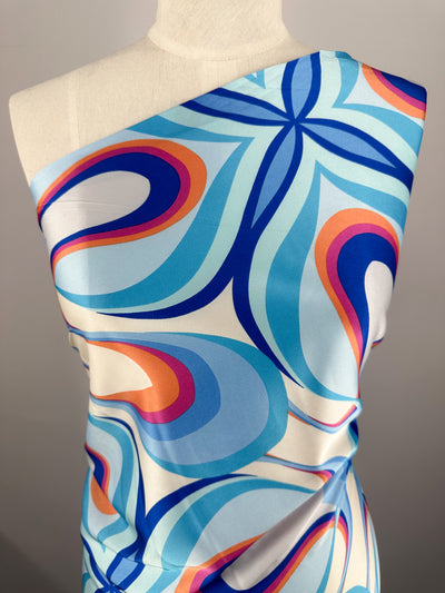 A mannequin showcases the vibrant Silky Satin - Burst - 150cm by Super Cheap Fabrics, featuring bold retro swirling shapes in blue, orange, and pink on lightweight satin for an eye-catching geometric effect.