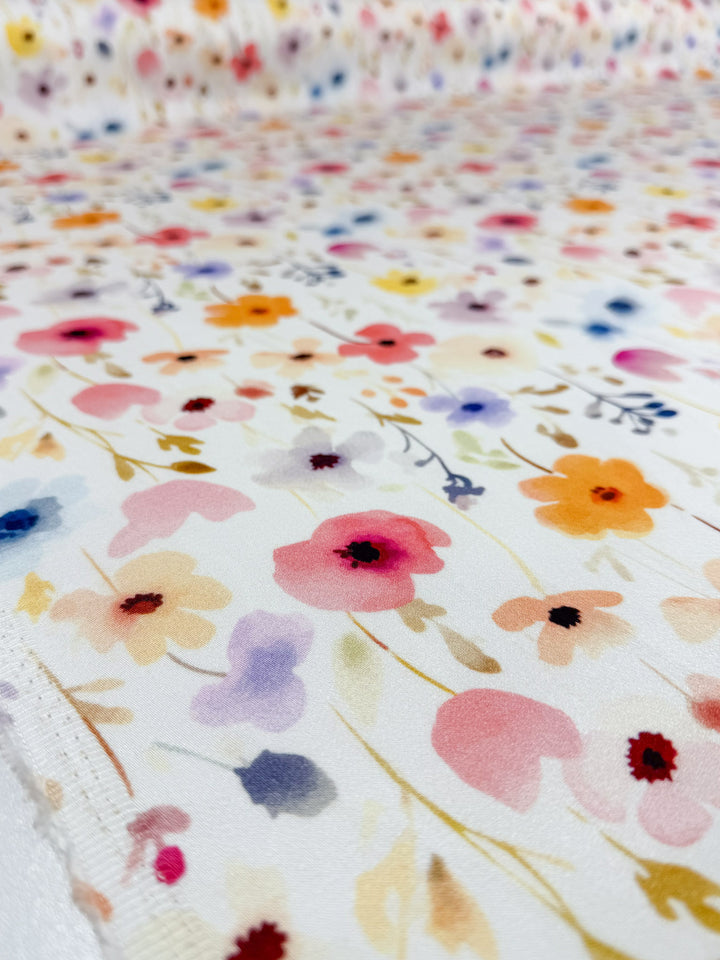 A close-up of Super Cheap Fabrics' Mercerised Rayon - Meadowdrop (150cm) shows lightweight white fabric with a colorful scattered floral pattern in pink, orange, purple, and blue, creating a soft watercolor effect and highlighting its detailed design.