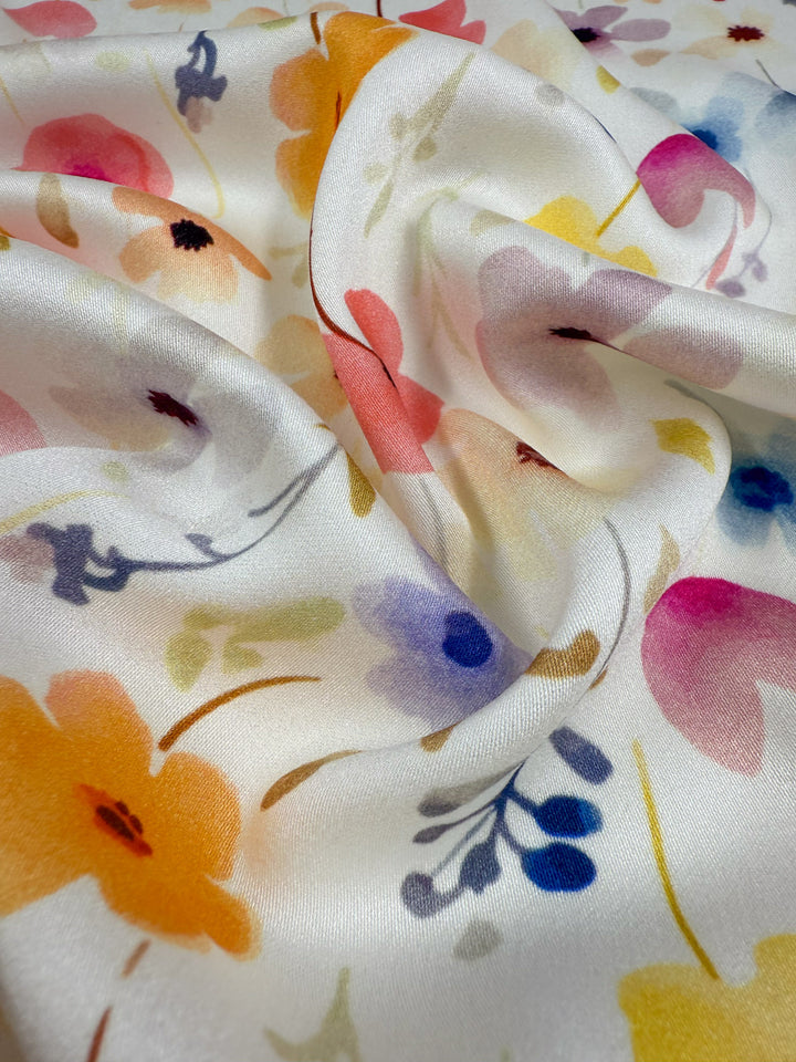 A close-up of Mercerised Rayon - Meadowdrop by Super Cheap Fabrics, a silky white lightweight fabric with orange, pink, blue, and yellow watercolor-style flowers, softly draped with smooth folds and a subtle sheen.