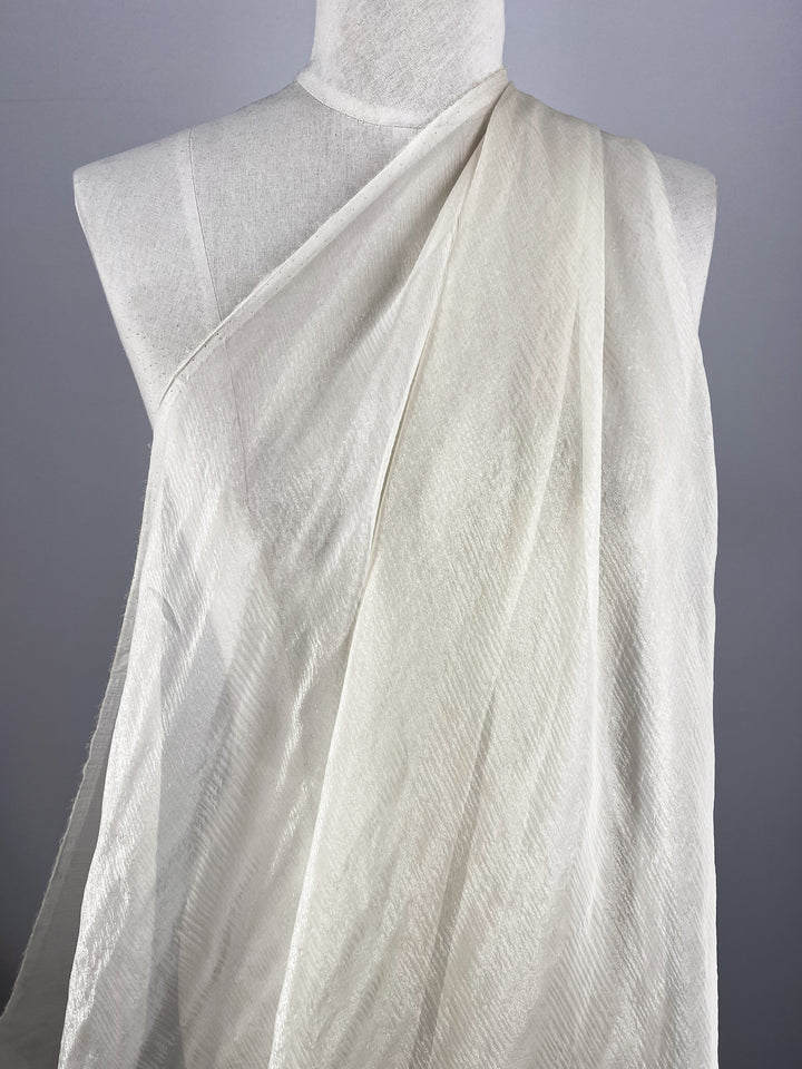 A white mannequin showcases Super Cheap Fabrics' Silk Mousseline in Milk, lightly draped over one shoulder for a flowing look with a subtle metallic shimmer against a plain gray backdrop.