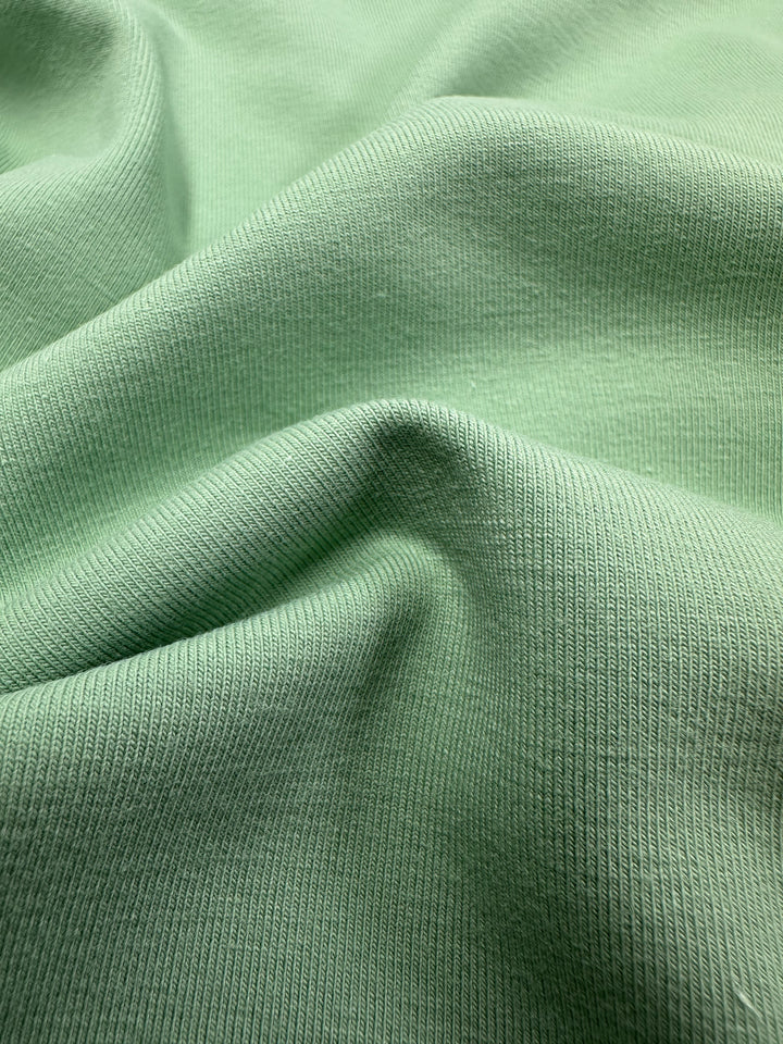 Close-up of Super Cheap Fabrics’ Cotton Lycra - Spruce Stone (170cm) in light green, featuring a ribbed texture, gentle folds, and a soft, smooth weave.