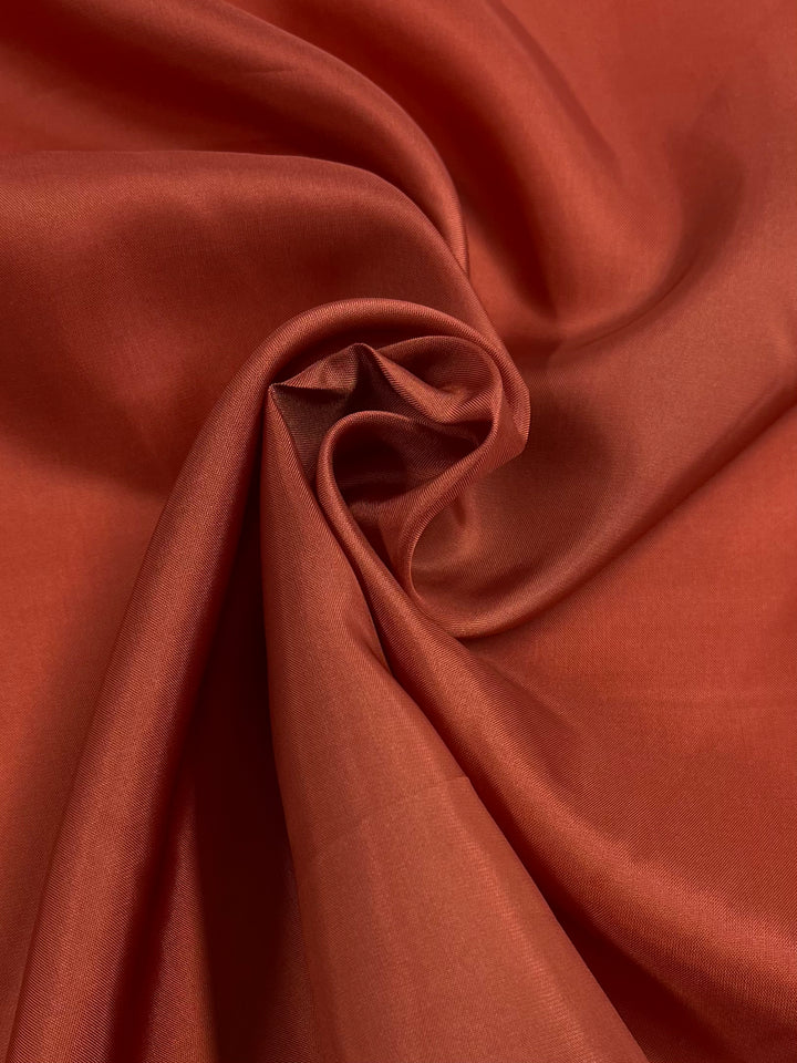 Close-up of rust-colored fabric. The lightweight material is gathered and twisted into a rosette shape in the center, creating soft folds and creases that showcase its smooth texture. The lighting highlights the fabric's satin-like sheen, typical of Super Cheap Fabrics' Lining - Burnt Ochre - 140cm.