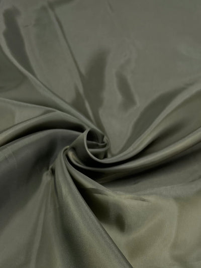 A close-up image of a piece of shiny, smooth, olive-green lightweight fabric. The material is gently gathered and twisted in the center, creating soft folds and highlights that catch the light, giving the Super Cheap Fabrics Lining - Olive - 112cm a glossy appearance.