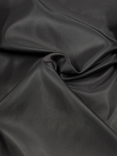 A close-up image of smooth, folded charcoal fabric with a satin-like sheen. This 100% polyester light-weight fabric appears slightly glossy, with soft creases and gentle curves adding texture to the material. The product is Lining - Charcoal - 120cm by Super Cheap Fabrics.