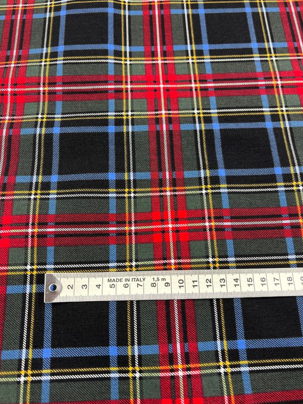 A close-up of the vibrant Printed Bengaline - Uniform fabric by Super Cheap Fabrics, featuring a tartan pattern in red, green, black, blue, and yellow crisscross stripes. A centimeter measuring tape lay across the 150cm-wide fabric to show scale.
