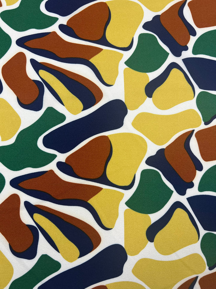 The Printed Crepe - Camo by Super Cheap Fabrics features an abstract pattern of green, brown, yellow, and navy shapes on white. Its design evokes scattered leaves and organic forms, ideal for high-fashion pieces crafted from lightweight fabric. Width: 150cm.
