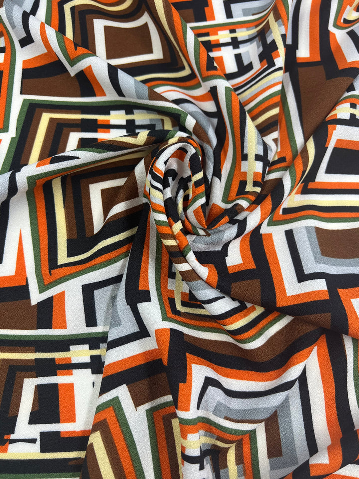 A close-up of the Printed Crepe - Geo Repeat by Super Cheap Fabrics, showcasing colorful polyester fabric with swirling geometric patterns. The design includes overlapping squares and rectangles in orange, brown, green, white, and black for a dynamic look ideal for high fashion.