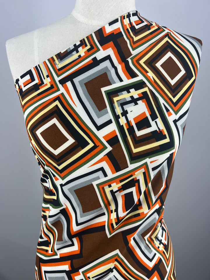 A mannequin displays Printed Crepe - Geo Repeat fabric by Super Cheap Fabrics, showcasing a retro geometric pattern with overlapping squares and rectangles in brown, orange, green, and white on a plain gray background.